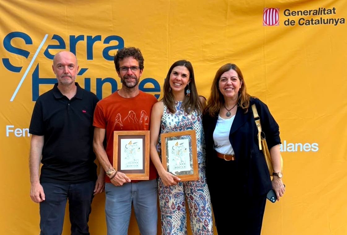 Quatre persones posen davant del photocall Serra Húnter i l’emblema de la Generalitat de Catalunya. Dues de les persones situades al centre sostenen marcs de fusta amb un reconeixement de l’esdeveniment.