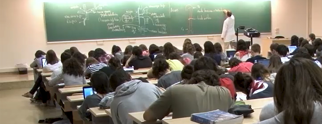  Vista general d'una aula amb alumnes. Al fons es veu un professor escrivint a la pissarra.