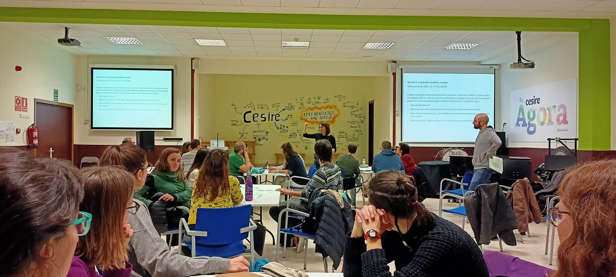 Aula amb diverses persones assegudes en grups escoltant una presentació, amb dues pantalles projectades i una professora explicant continguts davant d’una paret amb esquemes durant en el curs 