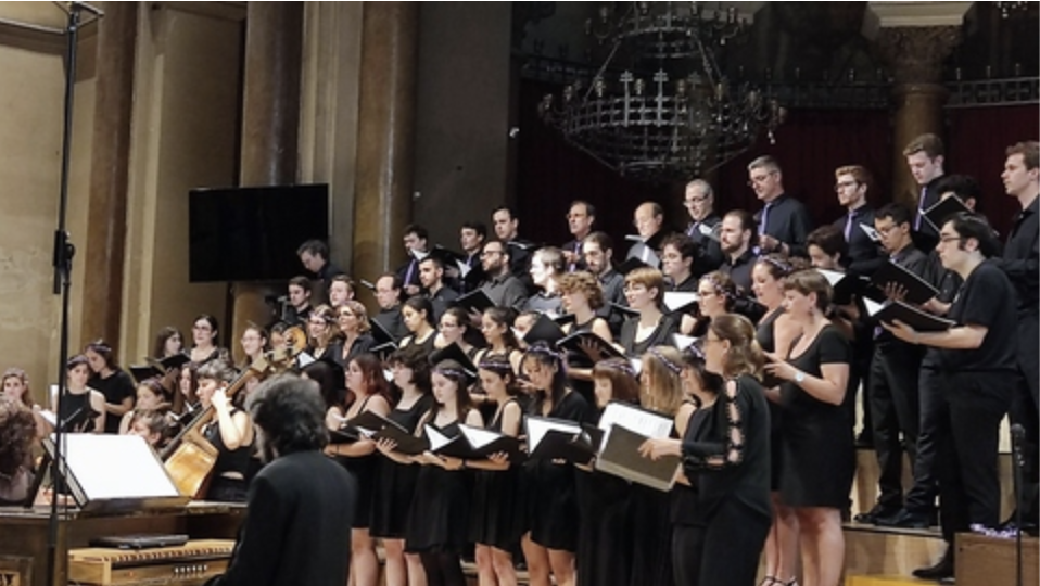    Cor i orquestra de la UAB interpretant una peça musical en un escenari interior, amb cantaires vestits de negre i un director dirigint la interpretació