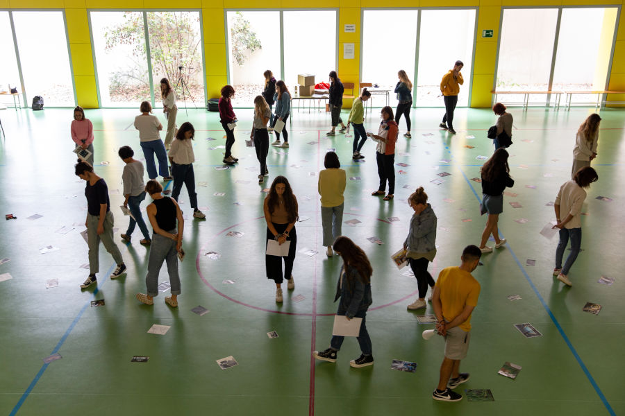  Grup de persones distribuïdes en un espai interior ampli, caminant i observant imatges col·locades a terra, en una activitat artística o educativa participativa.