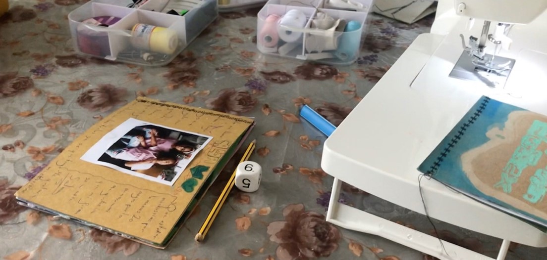  Taula de treball amb una màquina de cosir, caixes amb materials de costura, una llibreta amb una fotografia, un llapis i un dau, en un espai creatiu i artesanal.