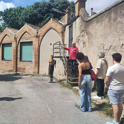 Amplia la imatge Grup escoltant una explicació davant d'edificis de maó amb arcs, amb una persona parlant des d'una escala