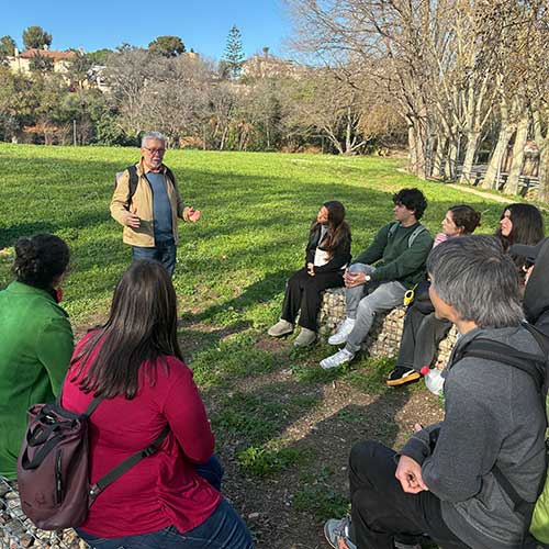 Amplia la imatge Grup escoltant una explicació en un parc, assegut i dret, al costat d'un cartell i zones verdes
