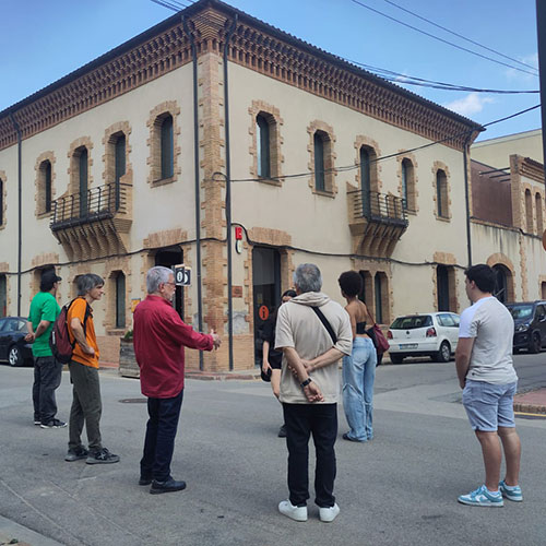 Amplia la imatge Grup de persones escoltant una explicació en una cruïlla, davant d'un edifici amb balcó i façana clara amb detalls de maó