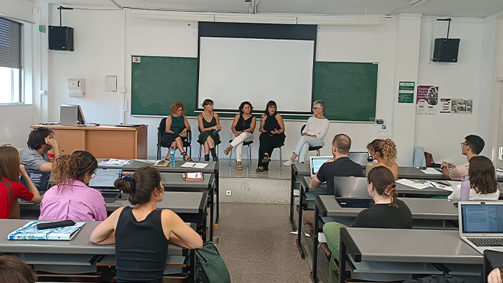 In a classroom with several rows of desks, a group of students is seated in front of four women and a person who appears to be the moderator, positioned on the stage in front of a projection screen. The speakers are talking.
