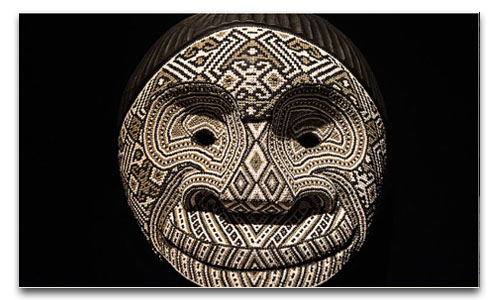 Traditional mask decorated with intricate geometric patterns in white, black and brown tones, set against a black background.