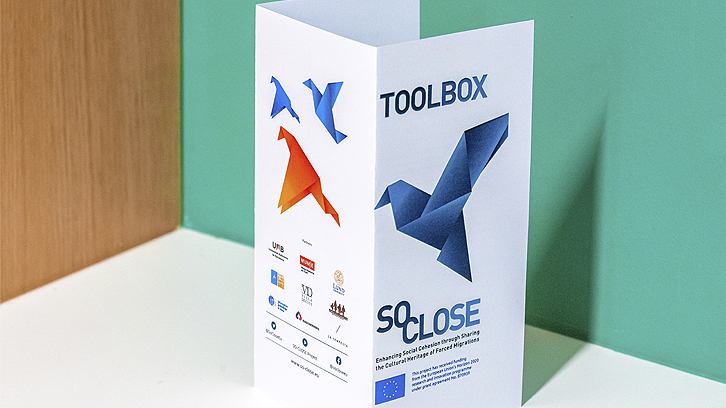 A tri-fold brochure is placed on a white surface, with a green wall in the background. The brochure features origami-style bird figures in blue and orange, with the text