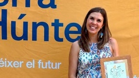 Una mujer que aparece de pie a la derecha de la imagen sostiene un marco de madera en el que se ve una ilustración y el nombre del evento.